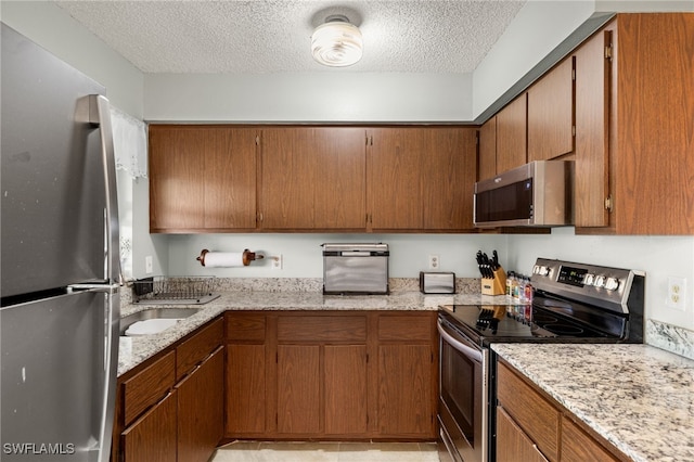 kitchen with a textured ceiling, light stone counters, stainless steel appliances, a sink, and brown cabinetry