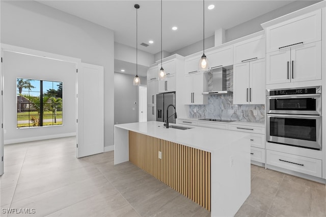 kitchen featuring a kitchen island with sink, a sink, tasteful backsplash, appliances with stainless steel finishes, and wall chimney exhaust hood