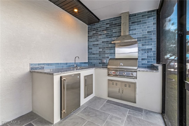 kitchen with a textured wall, high quality fridge, light stone countertops, wall chimney range hood, and a sink