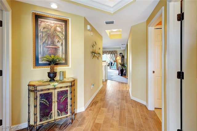 hallway featuring baseboards, visible vents, ornamental molding, a tray ceiling, and light wood-type flooring