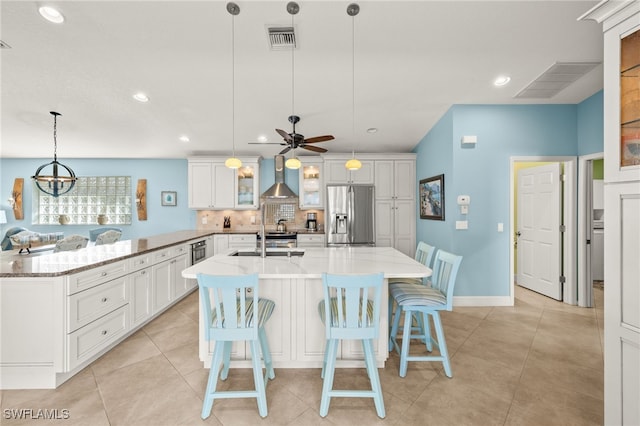 kitchen featuring visible vents, an island with sink, wall chimney exhaust hood, stainless steel refrigerator with ice dispenser, and backsplash