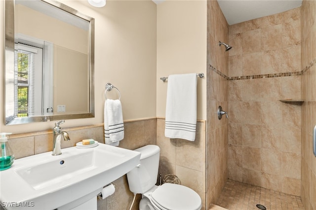 bathroom featuring toilet, a wainscoted wall, a sink, a tile shower, and tile walls