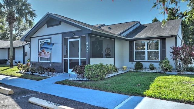 single story home with stucco siding, roof with shingles, a front yard, a sunroom, and brick siding