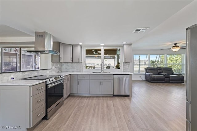 kitchen with stainless steel appliances, gray cabinetry, ceiling fan, and wall chimney range hood