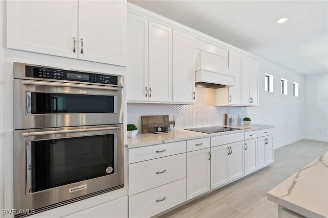 kitchen with black electric stovetop, stainless steel double oven, premium range hood, white cabinetry, and backsplash