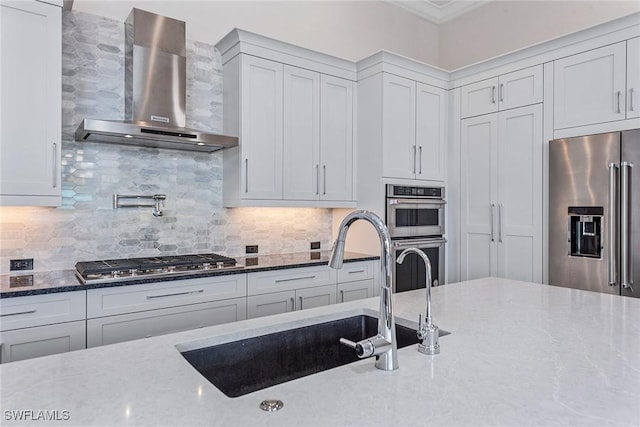 kitchen with light stone counters, a sink, backsplash, appliances with stainless steel finishes, and wall chimney exhaust hood