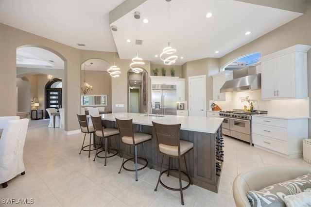 kitchen featuring high quality appliances, a large island with sink, light countertops, wall chimney exhaust hood, and a notable chandelier