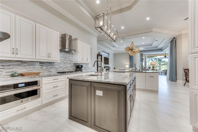 kitchen featuring a tray ceiling, a center island with sink, appliances with stainless steel finishes, a sink, and wall chimney exhaust hood