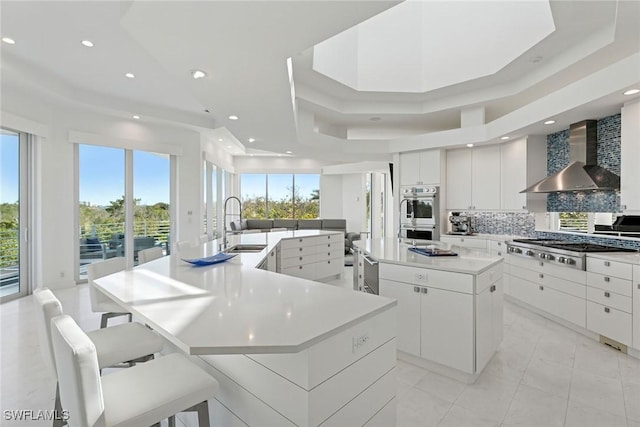 kitchen featuring stainless steel appliances, a raised ceiling, a large island with sink, and wall chimney range hood