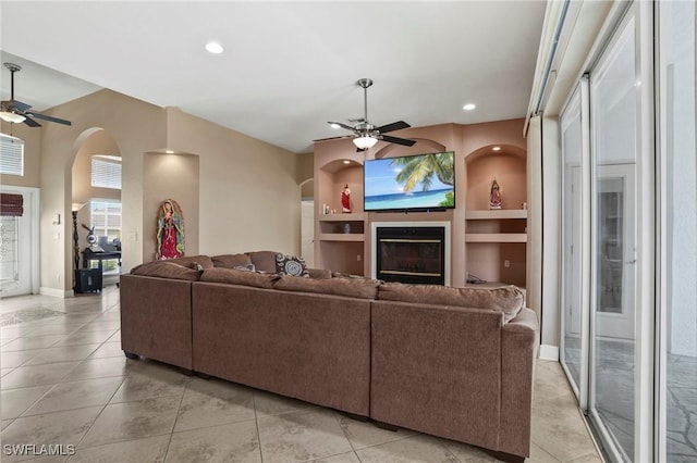 living room featuring a glass covered fireplace, built in shelves, arched walkways, and ceiling fan