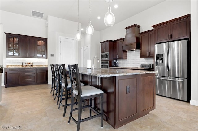 kitchen featuring custom range hood, a kitchen breakfast bar, a kitchen island with sink, stainless steel appliances, and dark brown cabinets