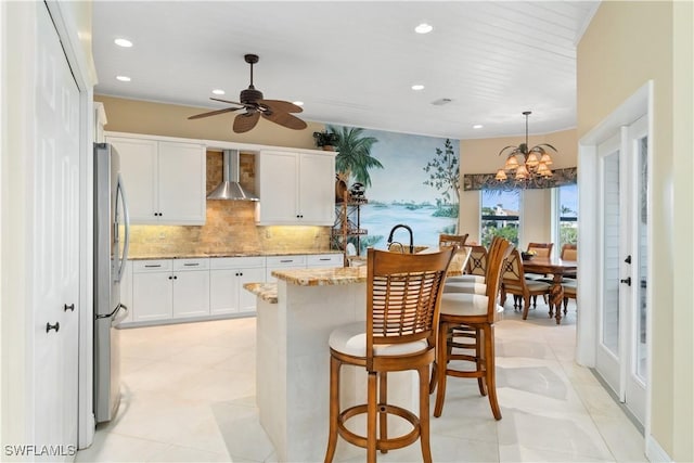 kitchen with wall chimney range hood, a breakfast bar, light stone counters, freestanding refrigerator, and white cabinets