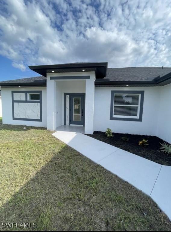 view of exterior entry with roof with shingles, a lawn, and stucco siding