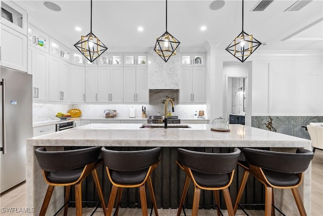 kitchen featuring light stone counters, a center island with sink, backsplash, light wood-style flooring, and stainless steel fridge