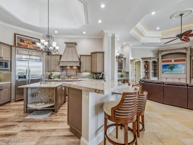 kitchen with backsplash, built in appliances, an island with sink, arched walkways, and a raised ceiling