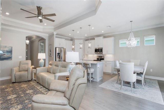 living room with light wood-type flooring, ornamental molding, ceiling fan with notable chandelier, arched walkways, and baseboards