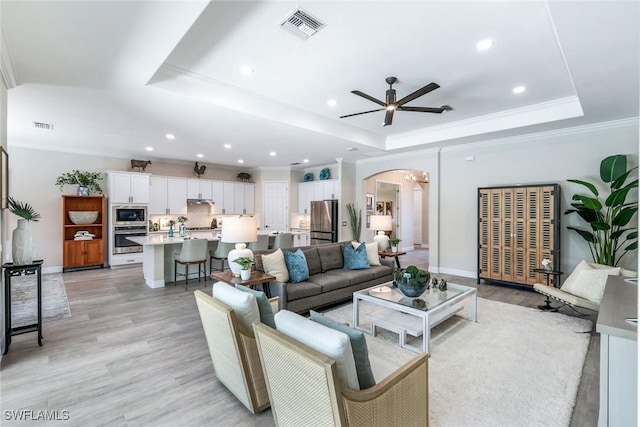 living area featuring visible vents, arched walkways, a raised ceiling, crown molding, and light wood-type flooring