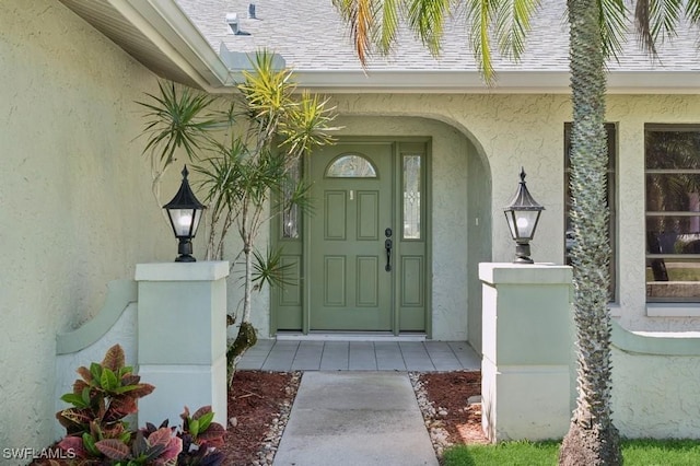 view of exterior entry with stucco siding and a shingled roof