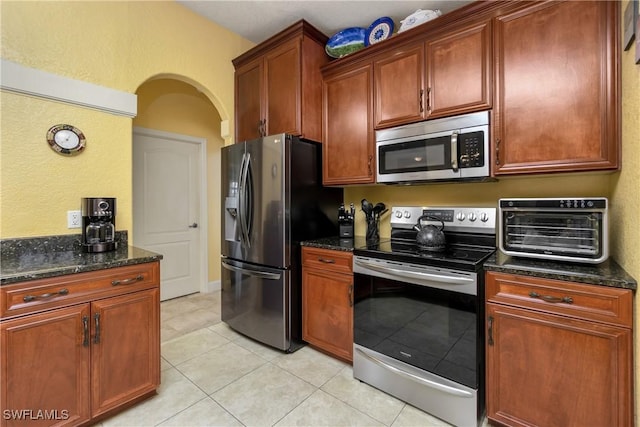 kitchen featuring a toaster, arched walkways, dark stone counters, stainless steel appliances, and light tile patterned flooring