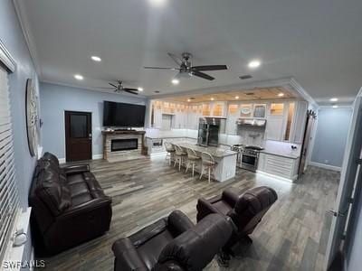 living area with a glass covered fireplace, ceiling fan, wood finished floors, crown molding, and recessed lighting