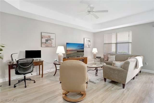 living room featuring light wood finished floors, ceiling fan, a raised ceiling, and baseboards