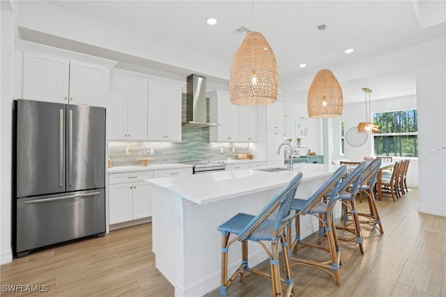 kitchen with white cabinets, light wood-style flooring, a kitchen island with sink, stainless steel appliances, and wall chimney range hood