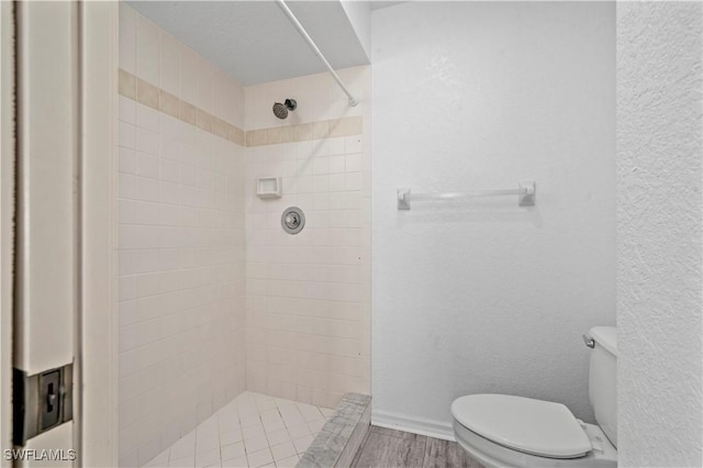 full bath featuring baseboards, toilet, wood finished floors, tiled shower, and a textured wall