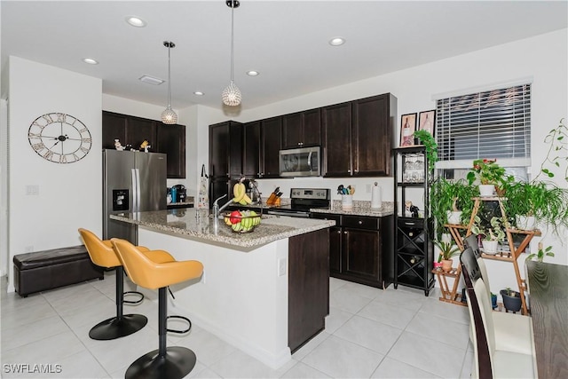 kitchen with light stone counters, a center island with sink, a breakfast bar, stainless steel appliances, and hanging light fixtures