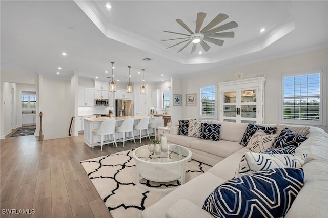 living room featuring visible vents, light wood-type flooring, a tray ceiling, ornamental molding, and a ceiling fan