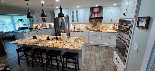 kitchen featuring a breakfast bar area, dark wood-type flooring, an island with sink, stainless steel fridge, and custom range hood