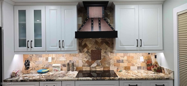 kitchen with tasteful backsplash, custom range hood, glass insert cabinets, white cabinetry, and black electric cooktop