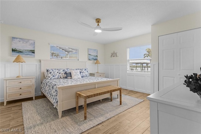 bedroom with a ceiling fan, light wood-style flooring, wainscoting, and a decorative wall