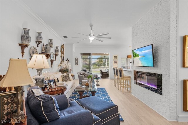 living area featuring visible vents, crown molding, light wood-style floors, a glass covered fireplace, and a ceiling fan