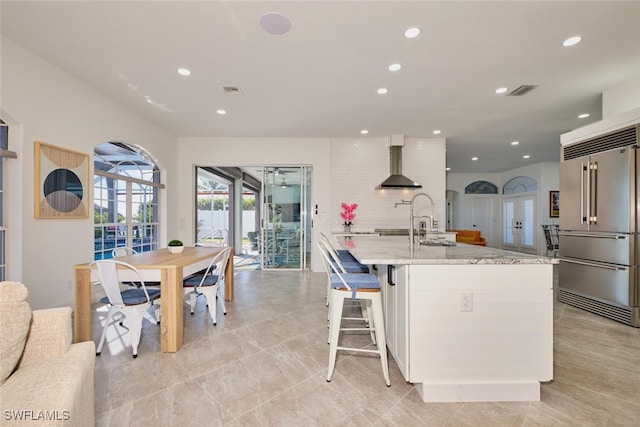 kitchen featuring a kitchen island with sink, light stone counters, recessed lighting, wall chimney exhaust hood, and high end refrigerator