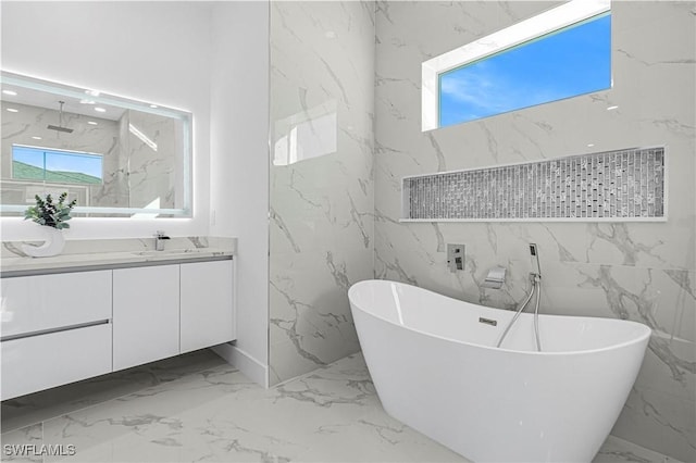 full bath featuring marble finish floor, a freestanding bath, vanity, and stone wall