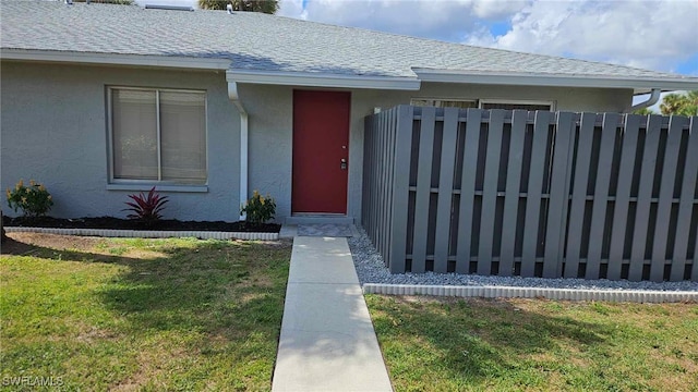 view of exterior entry with a shingled roof, a yard, fence, and stucco siding