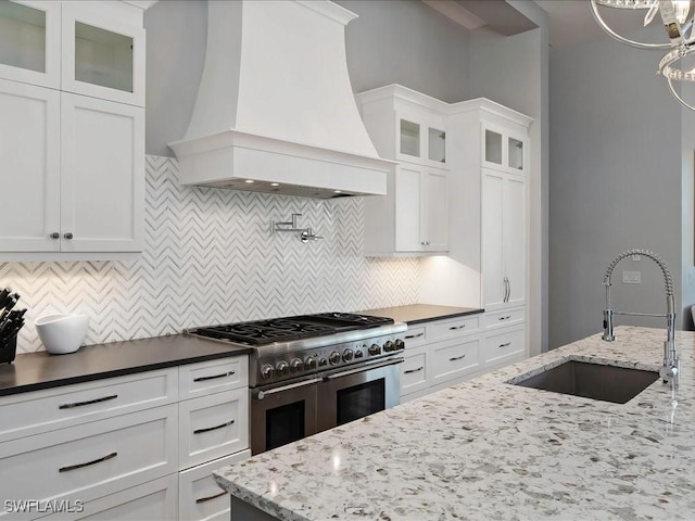 kitchen featuring tasteful backsplash, premium range hood, range with two ovens, white cabinets, and a sink