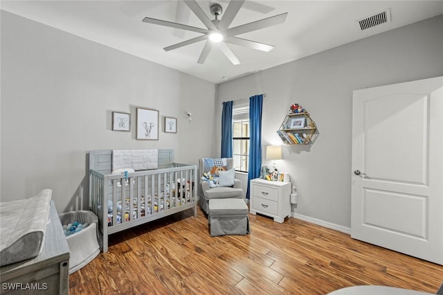 bedroom featuring wood finished floors, visible vents, baseboards, ceiling fan, and a nursery area