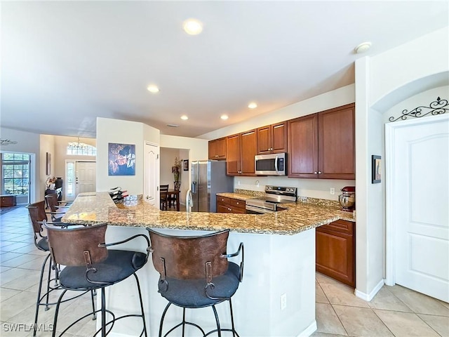 kitchen featuring a kitchen bar, light stone counters, appliances with stainless steel finishes, light tile patterned flooring, and brown cabinetry