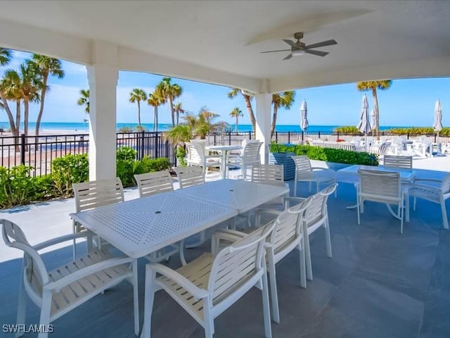 view of patio featuring a ceiling fan, fence, outdoor dining area, a water view, and a beach view