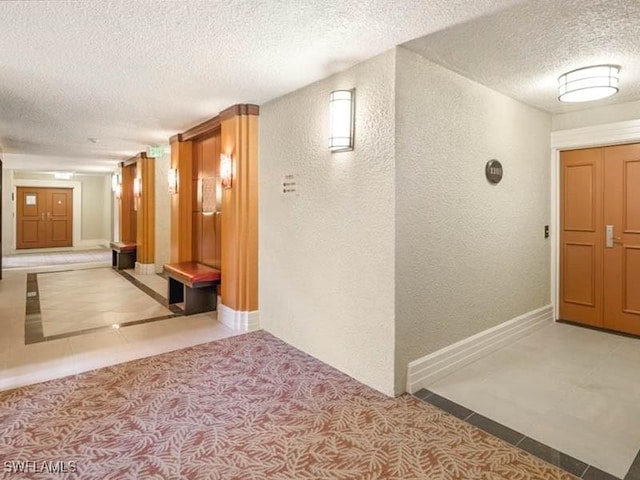 hallway featuring a textured wall, a textured ceiling, tile patterned flooring, and baseboards