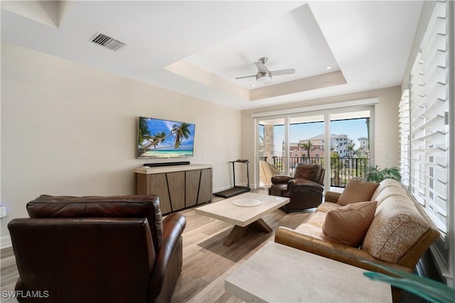 living room with light wood-type flooring, visible vents, baseboards, a raised ceiling, and ceiling fan