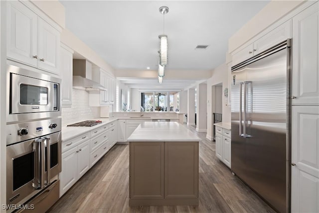 kitchen with a kitchen island, white cabinetry, wall chimney exhaust hood, light countertops, and built in appliances