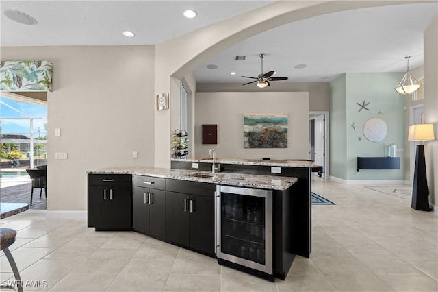 kitchen featuring beverage cooler, dark cabinets, arched walkways, a ceiling fan, and a sink