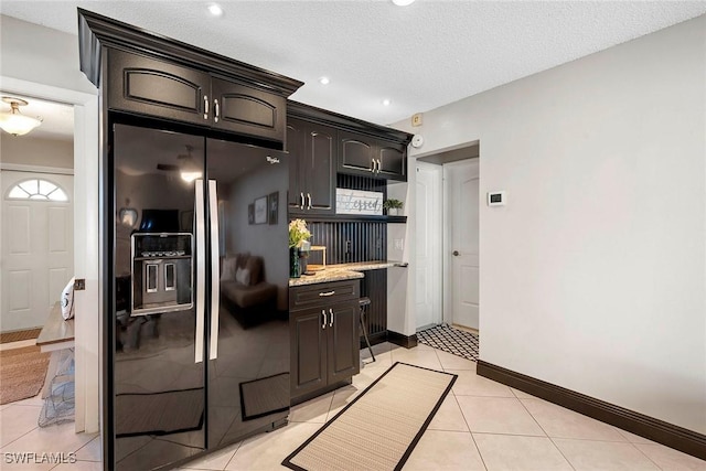 kitchen with light tile patterned floors, baseboards, dark brown cabinets, black refrigerator with ice dispenser, and light stone countertops