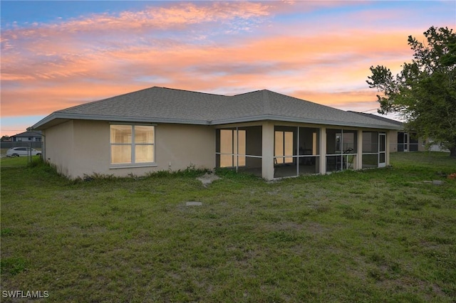 back of house at dusk featuring a shingled roof, a yard, a sunroom, and stucco siding