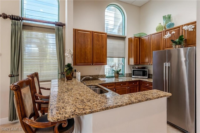 kitchen with a sink, stainless steel appliances, a peninsula, brown cabinetry, and light stone countertops