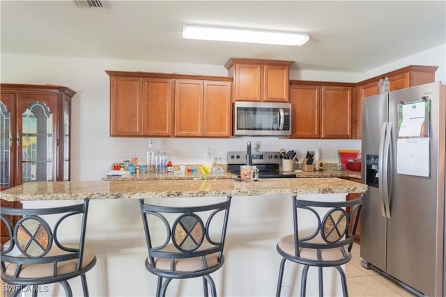 kitchen with light stone countertops, a center island with sink, light tile patterned flooring, stainless steel appliances, and brown cabinets