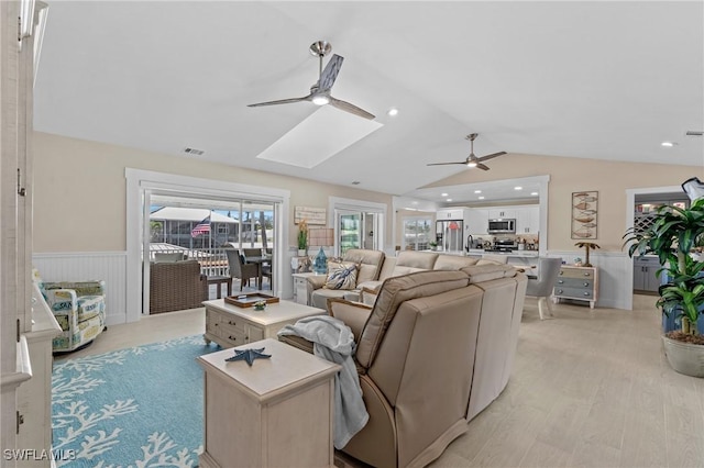 living room featuring a ceiling fan, visible vents, light wood finished floors, vaulted ceiling with skylight, and wainscoting