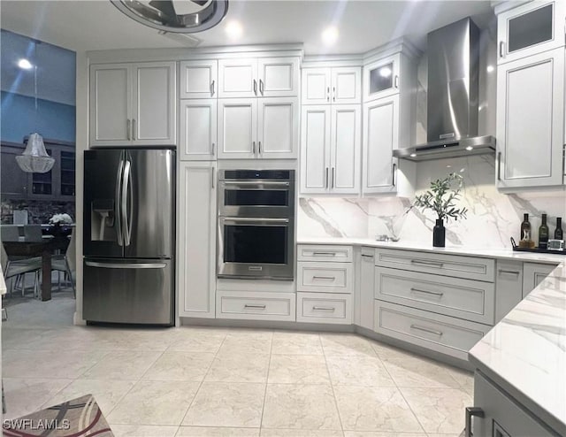 kitchen with glass insert cabinets, wall chimney range hood, light stone counters, decorative backsplash, and appliances with stainless steel finishes